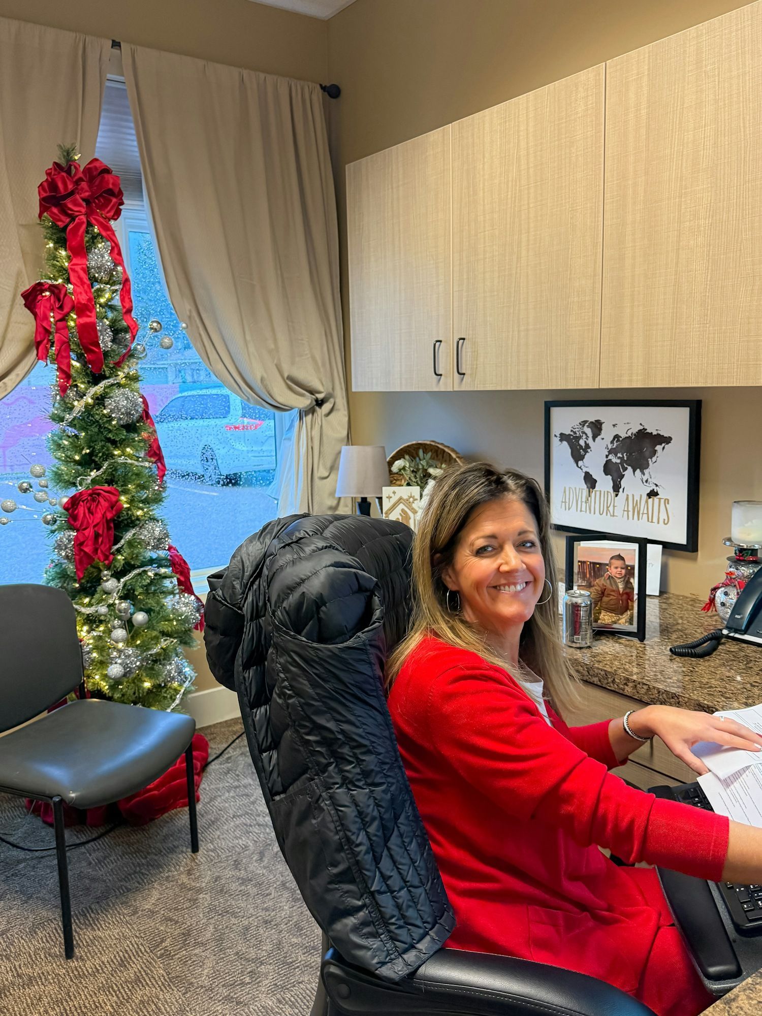 Woman in red sweater sits at desk, smiling. Christmas tree with red bows beside window.