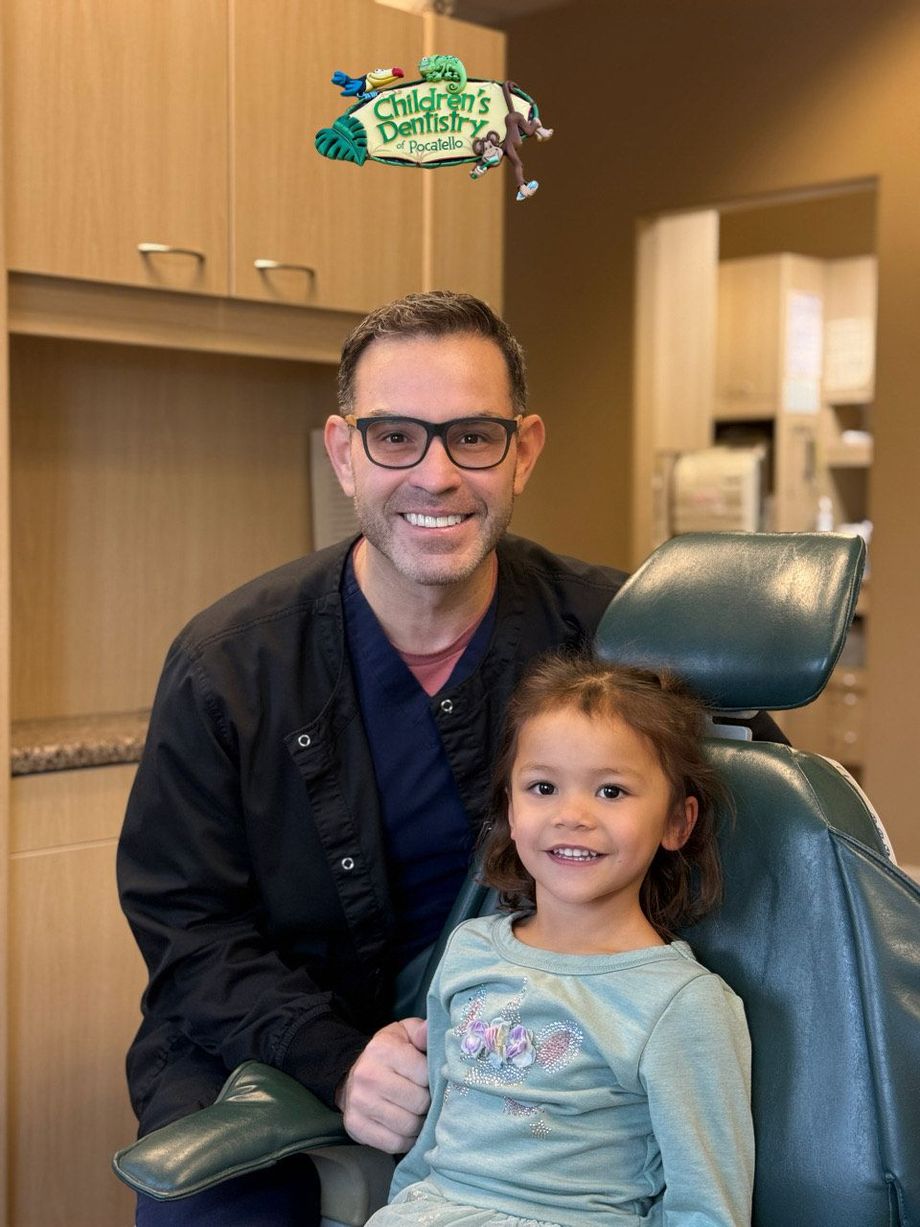 Dentist with smiling child in dental chair. Dentist wears glasses and scrubs. Light brown cabinetry in background.
