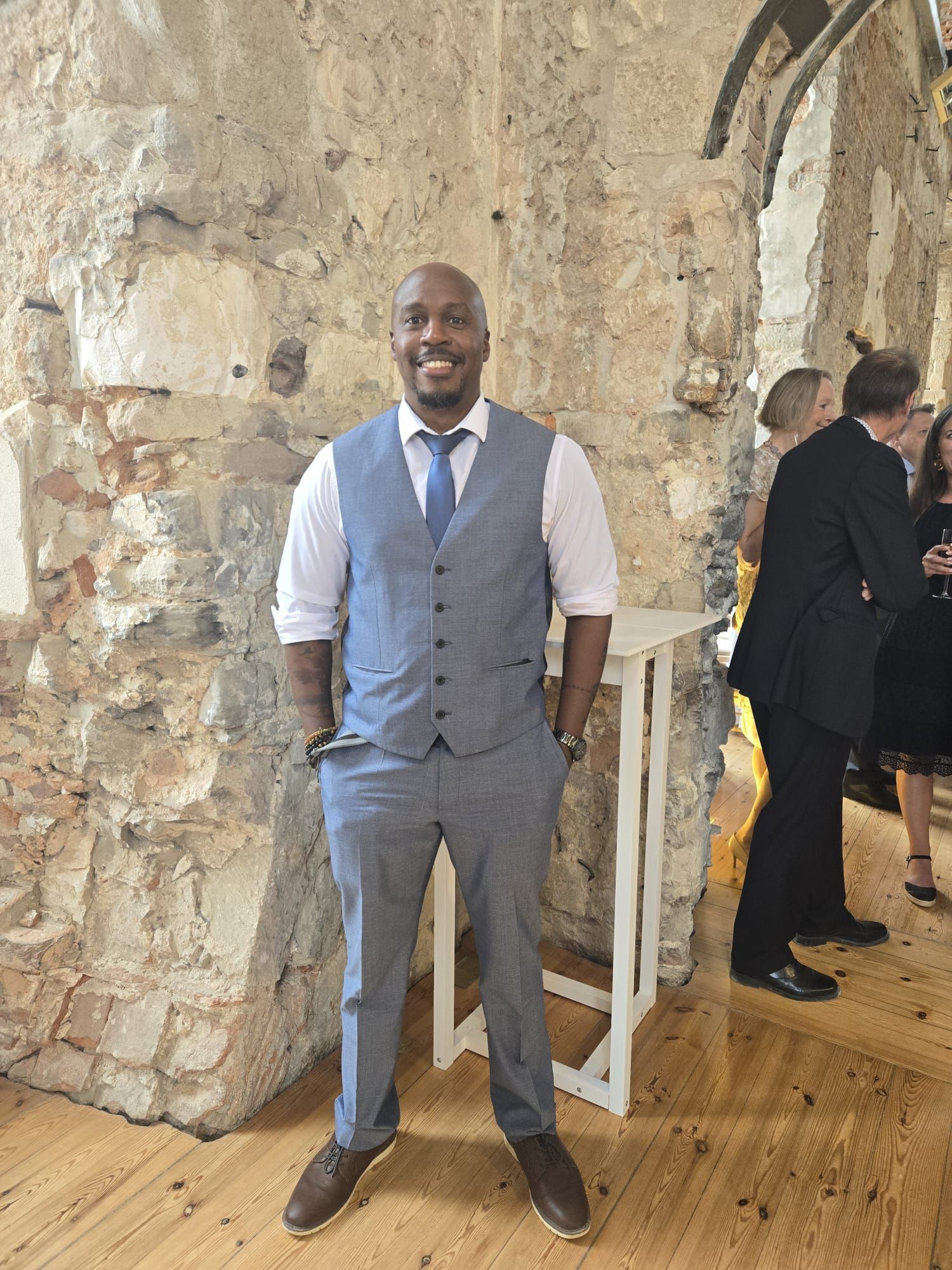 Man in blue suit, smiling, standing near white table, against stone wall.
