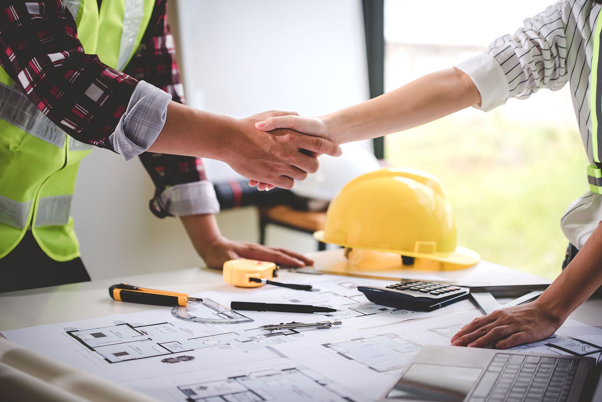 Two construction workers are shaking hands over a table.