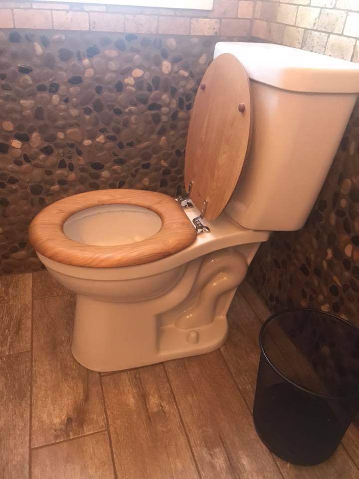 A toilet with a wooden seat is in a bathroom next to a trash can.