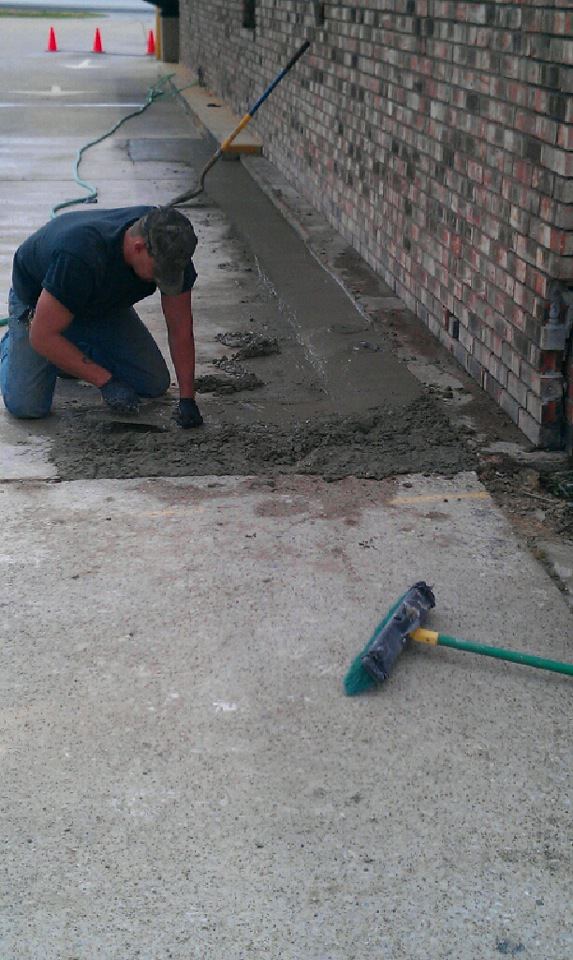 A man is kneeling on the ground spreading concrete next to a broom.