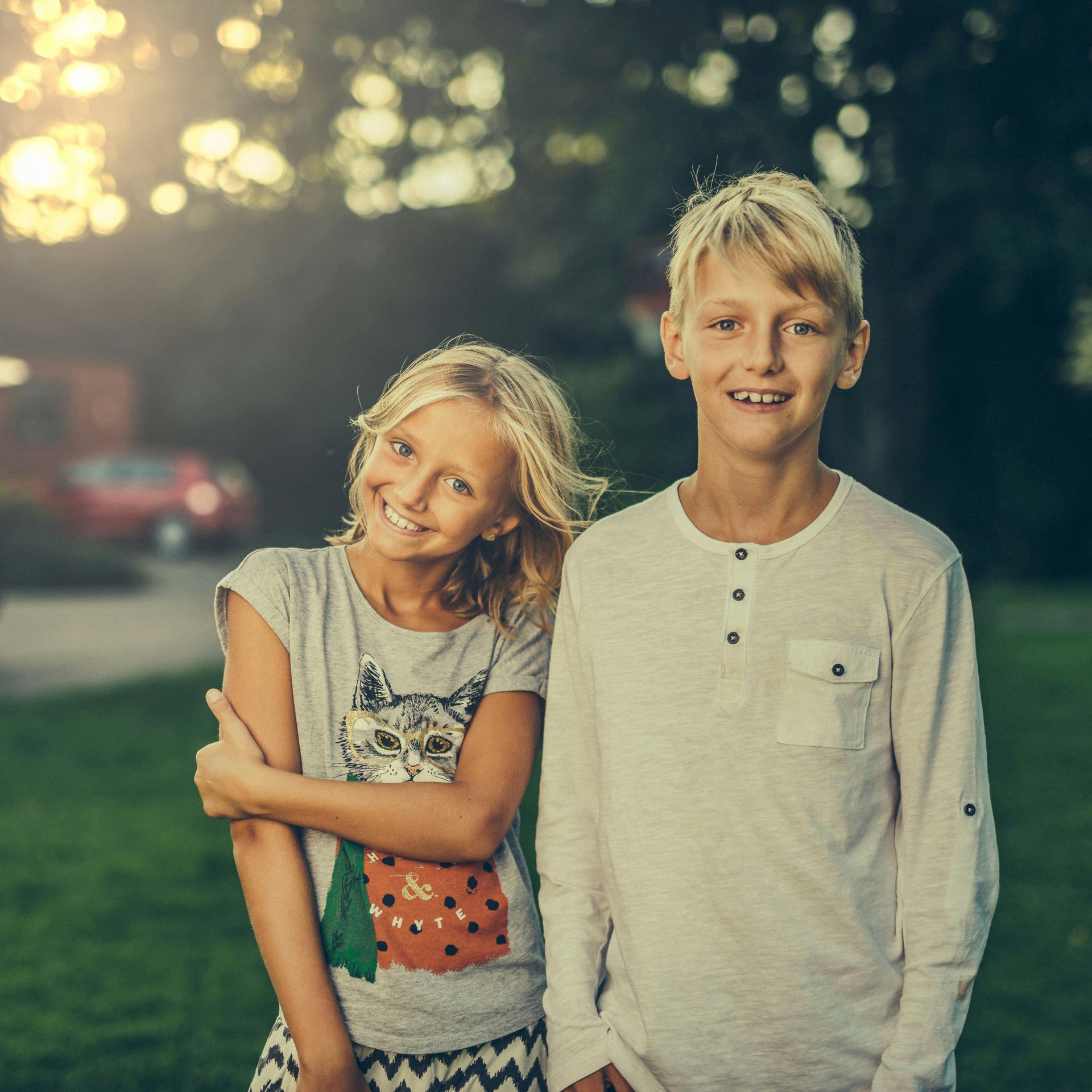 Two smiling children outdoors. Girl in a cat tee-shirt; boy in a long-sleeved shirt. Backlit by sun.