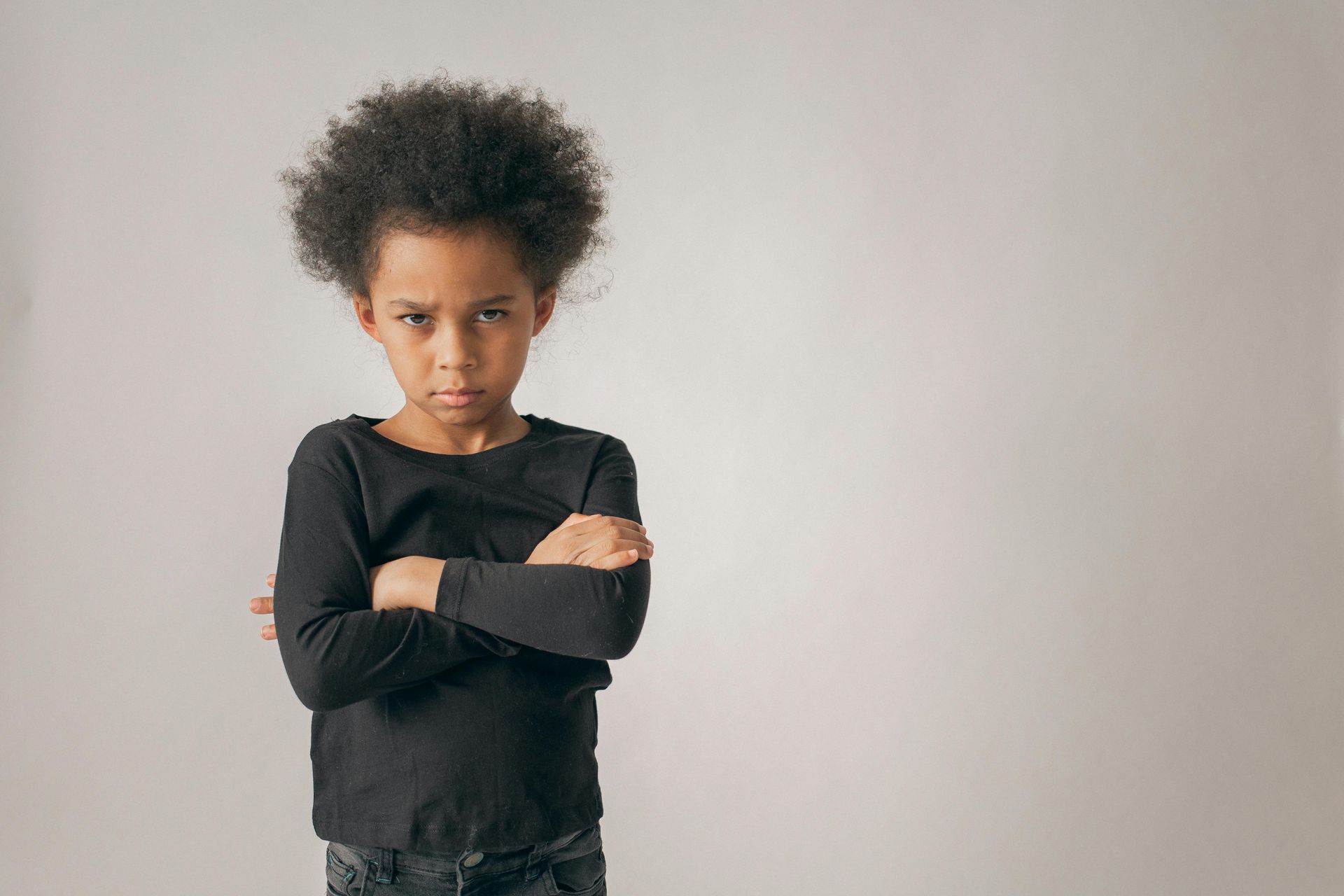 A child with crossed arms and a stern expression. They are wearing a black long-sleeved shirt, standing in front of a white backdrop.