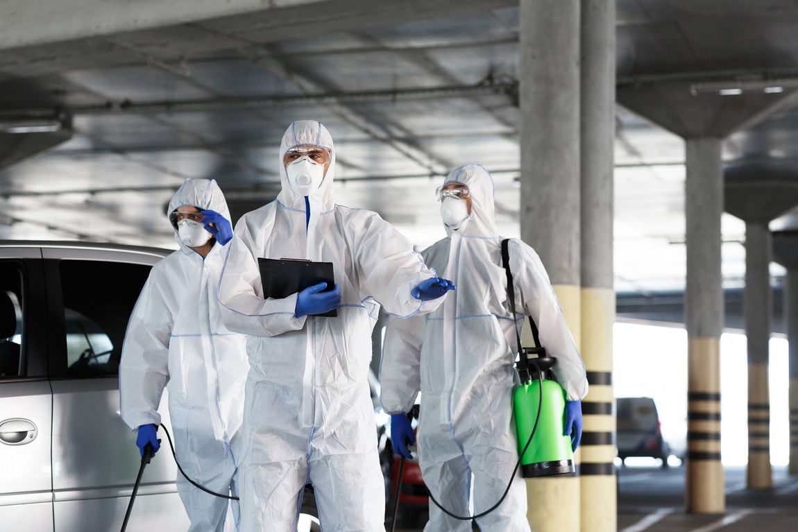 Three people in white hazmat suits inspect a parking garage. One holds a clipboard. Three people in white hazmat suits inspect a parking garage. One holds a clipboard.