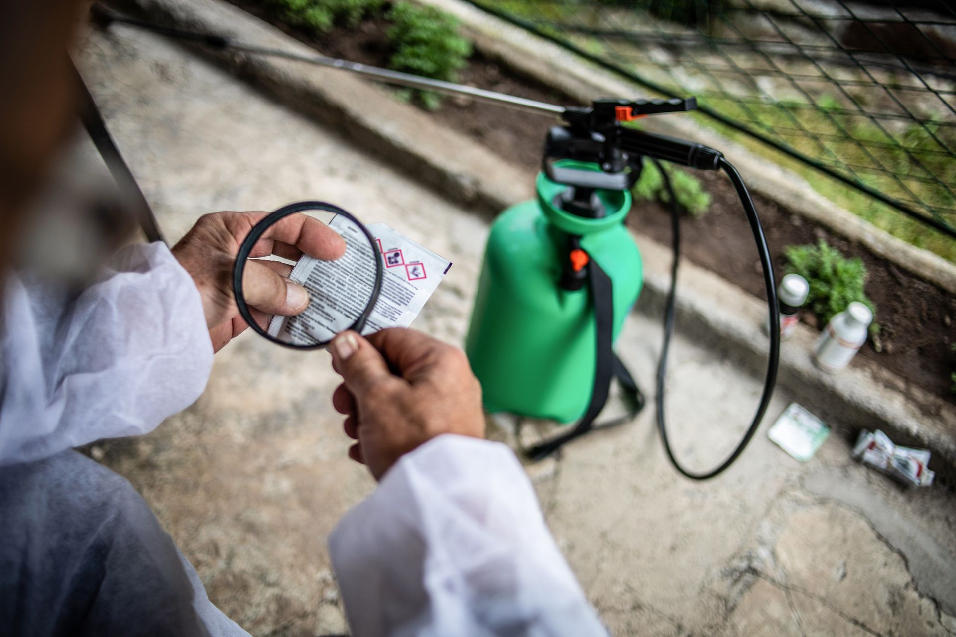 Person in protective suit examines a label with a magnifying glass, spray tank in background. Person in protective suit examines a label with a magnifying glass, spray tank in background.