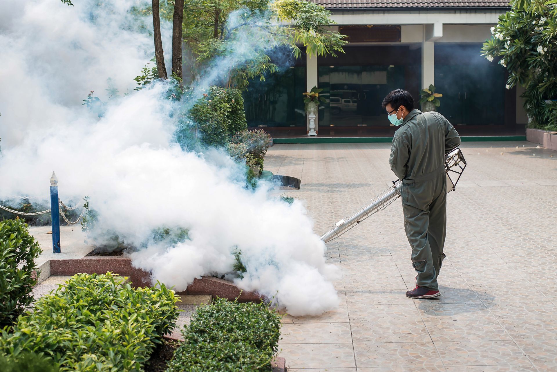 Man in protective suit sprays pesticide, creating fog in a paved area near foliage and a building.