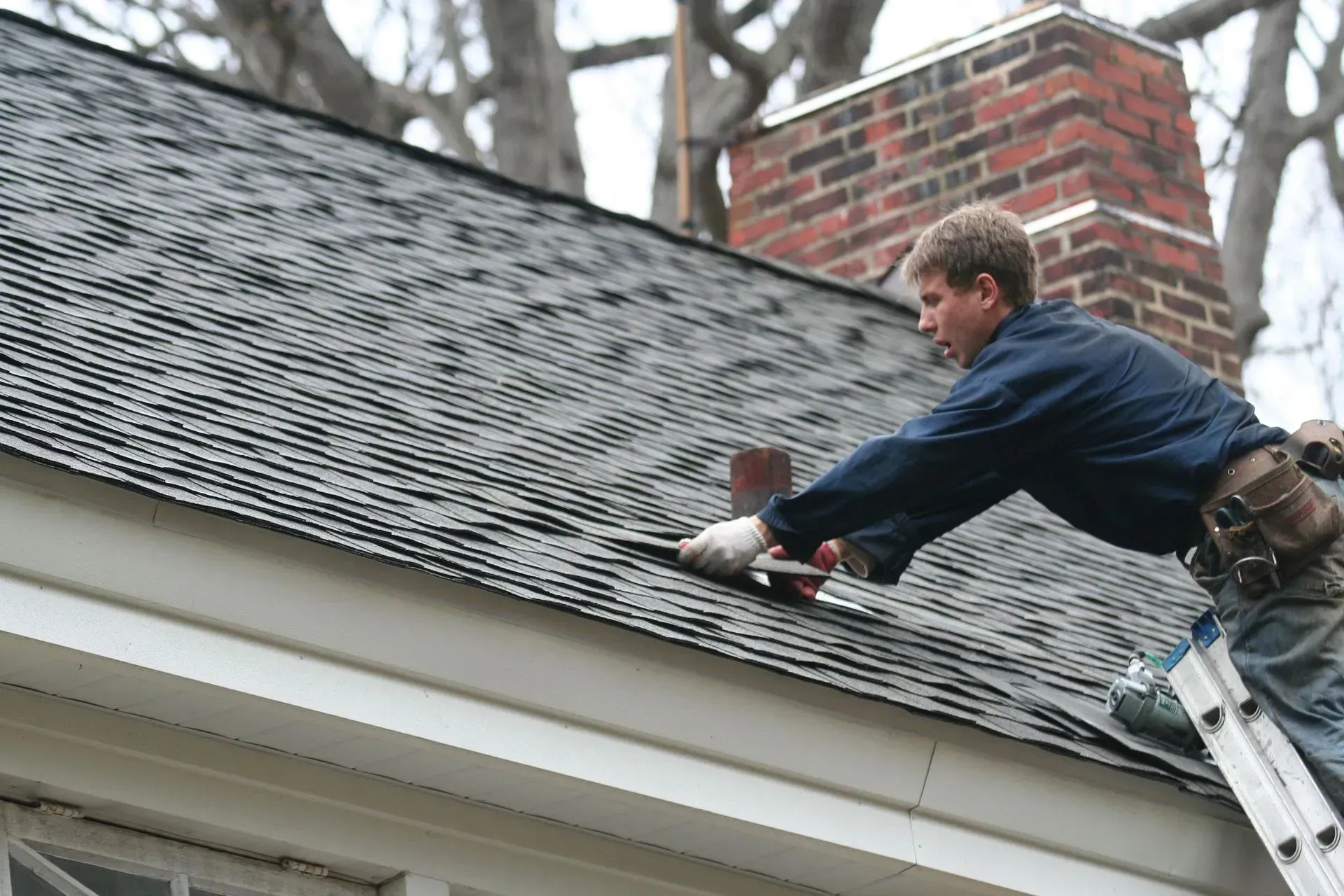 Roofer in blue jacket repairs shingles near a chimney, standing on a ladder.