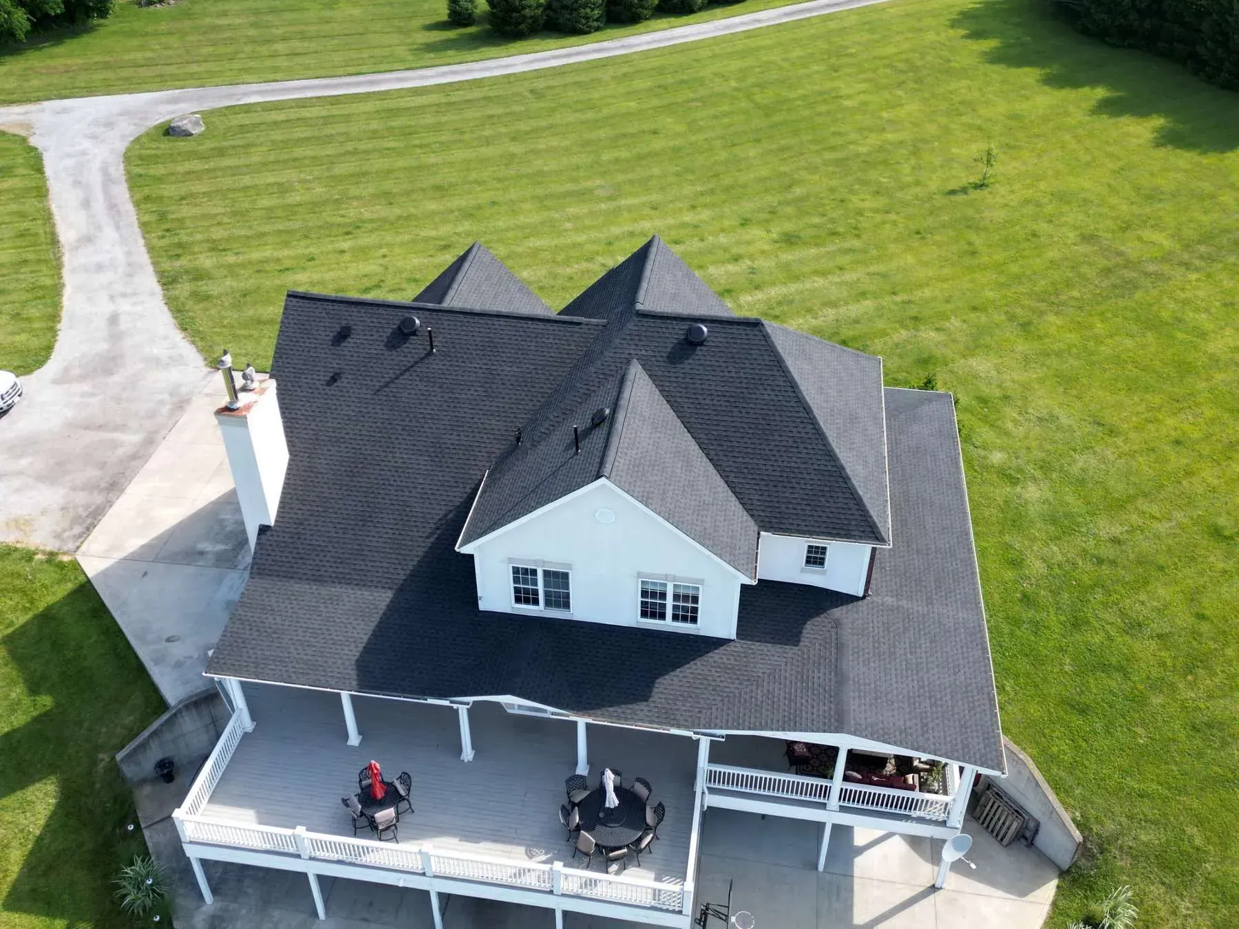 Aerial view of a white house with a dark roof and wraparound porch on a green lawn with a driveway.