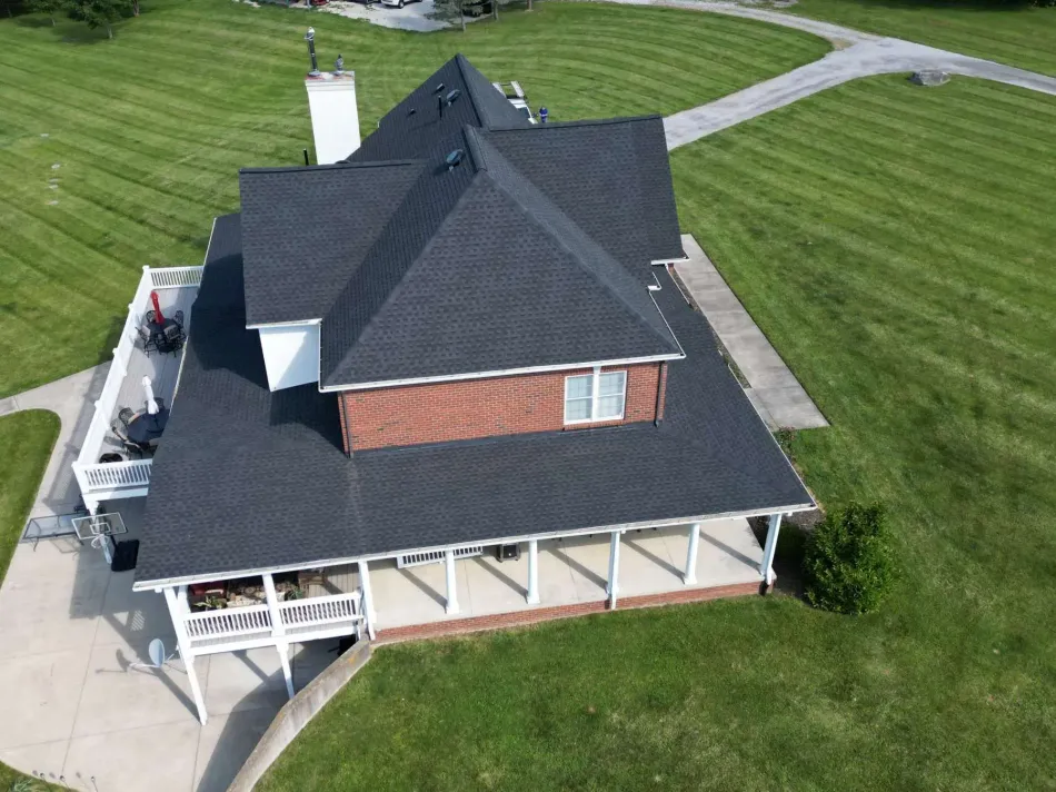 Person repairing shingle roof, holding a tool; white siding, brick chimney, and trees in background.