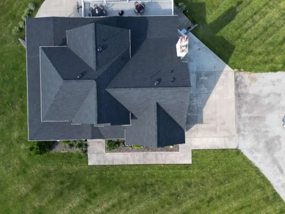 Overhead view of a dark shingled roof with a chimney, concrete driveway and lawn.