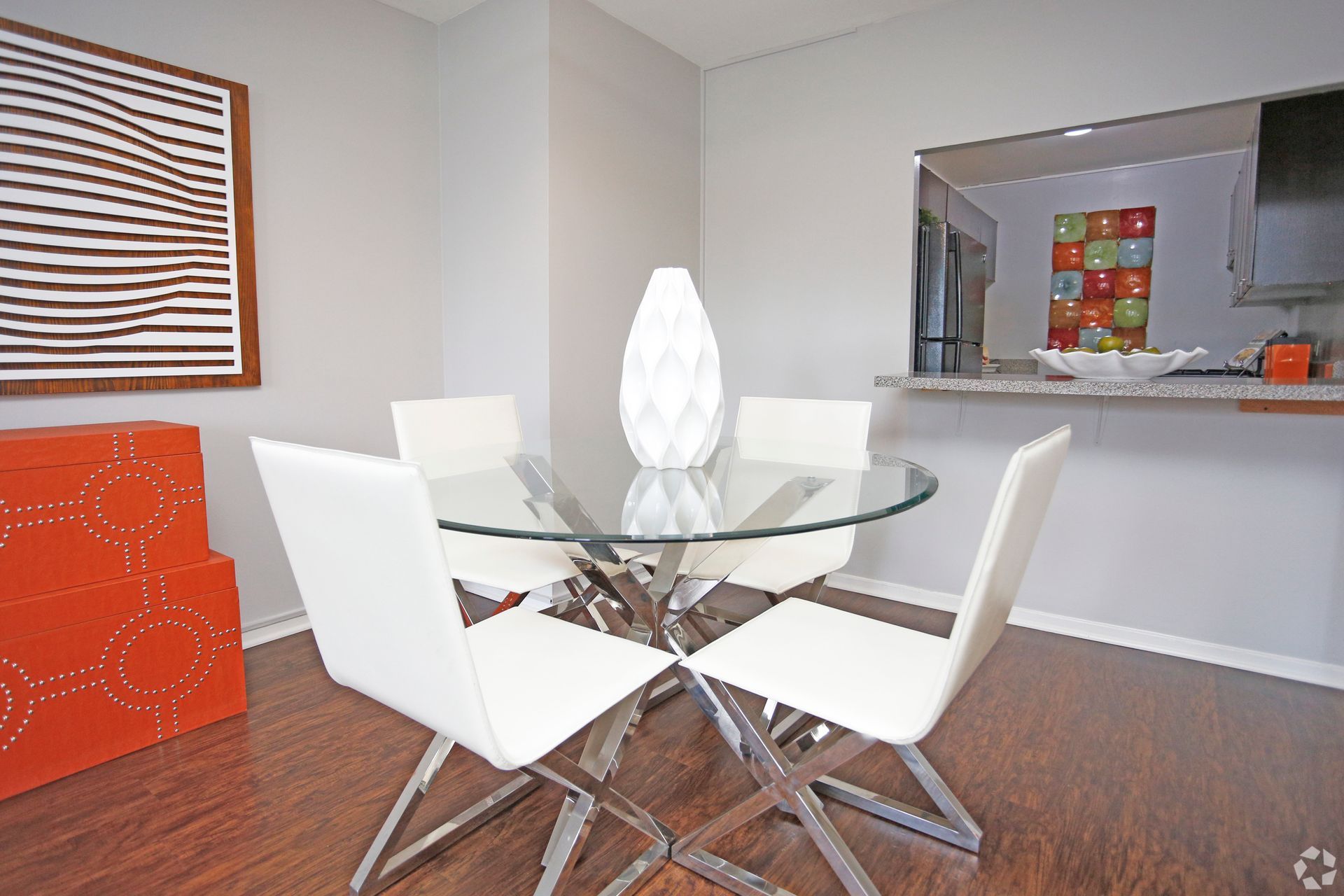 Dining room with glass table, white chairs, and a decorative vase. Kitchen visible in the background.