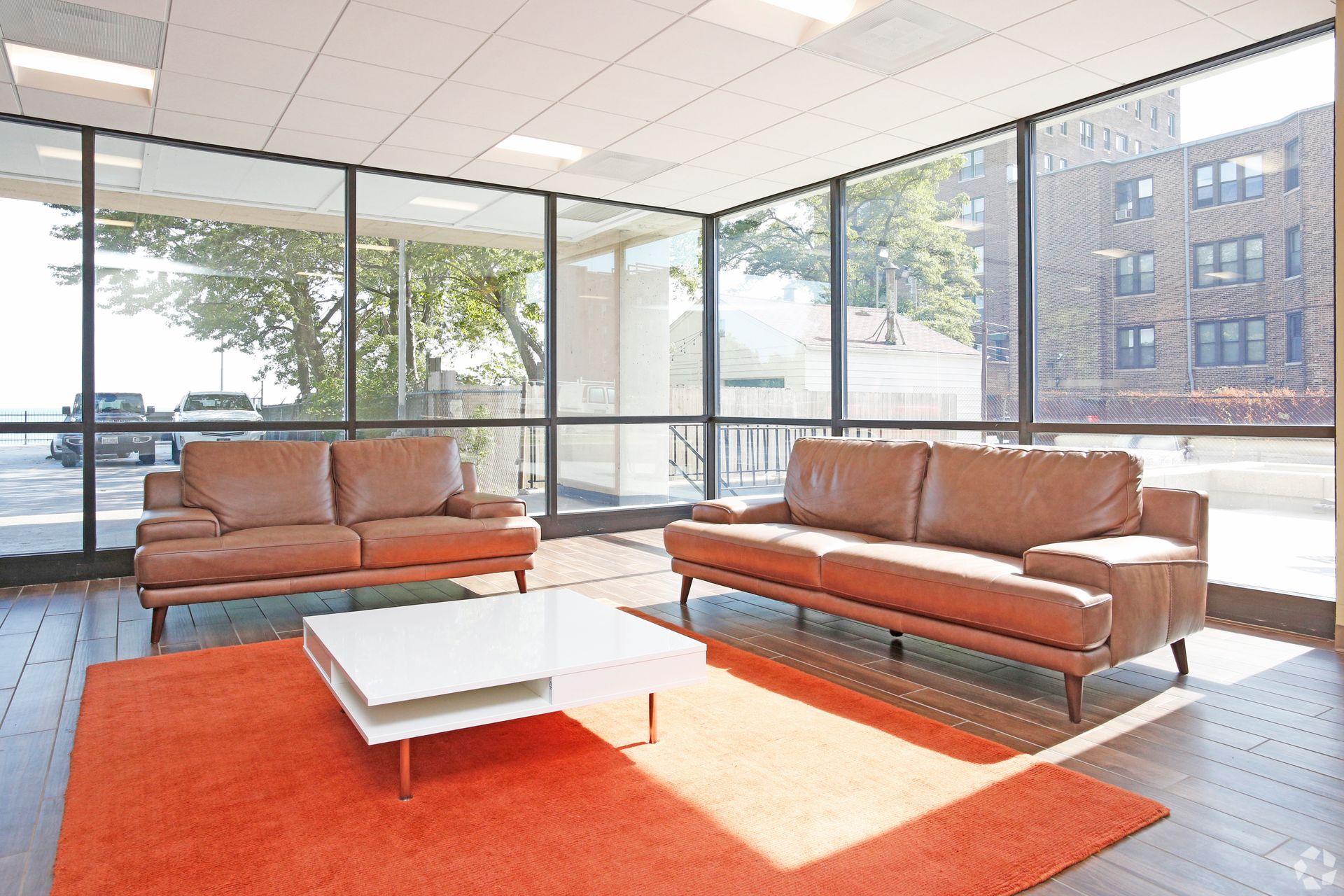 A lobby with brown leather couches, white coffee table, and orange rug; large windows offer outside views.