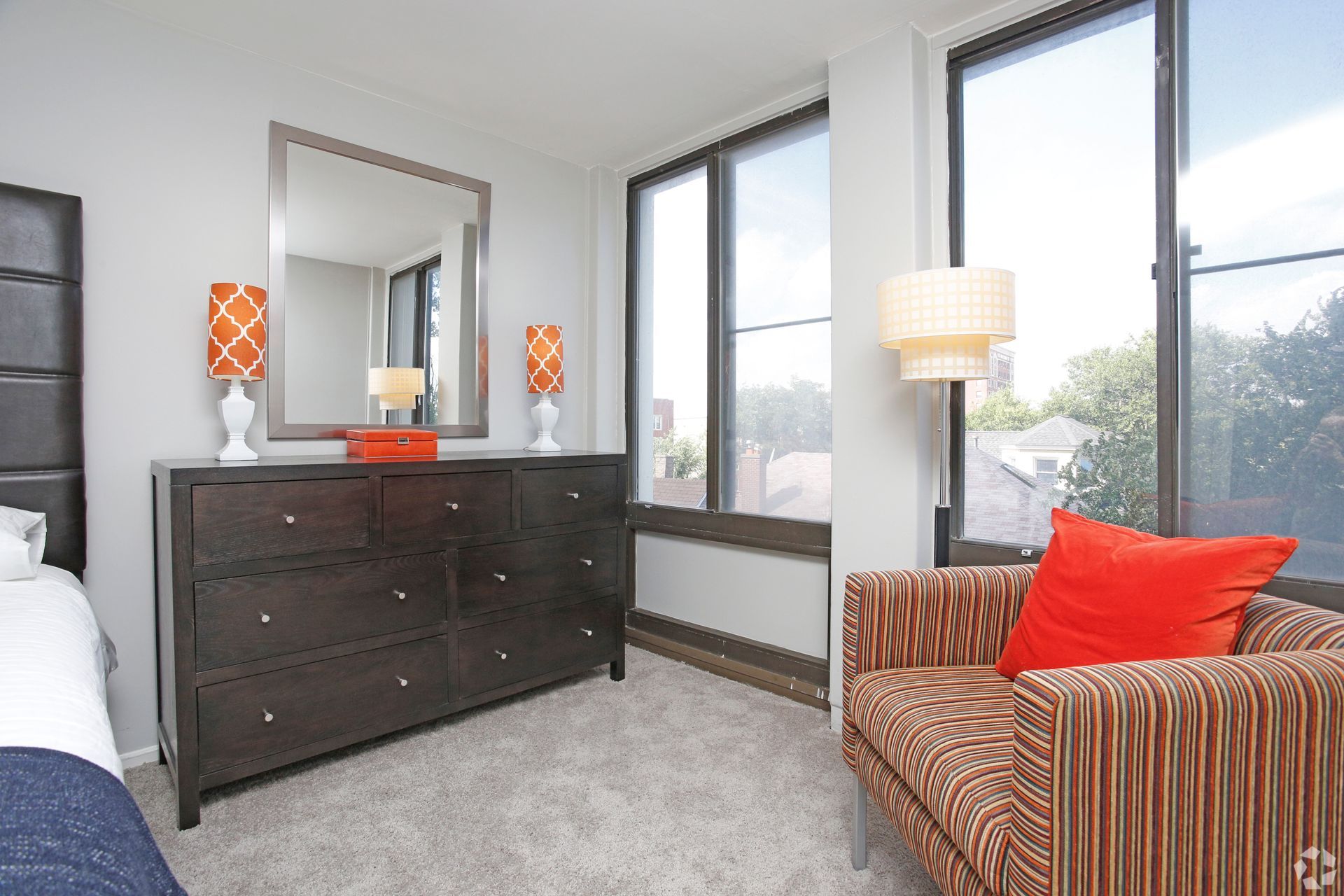 Bedroom with dark dresser, orange accents, armchair, and large windows.