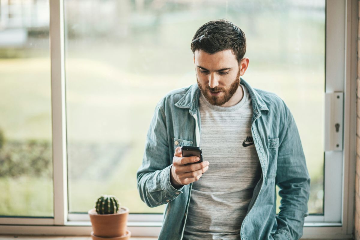 A man is standing in front of a window looking at his cell phone.