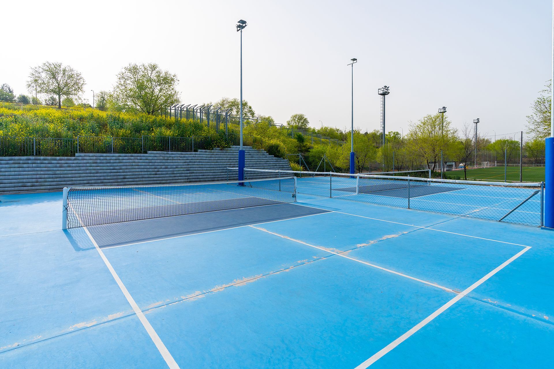 Blue pickleball court with net, lines, and light poles, outdoors near greenery and bleachers.