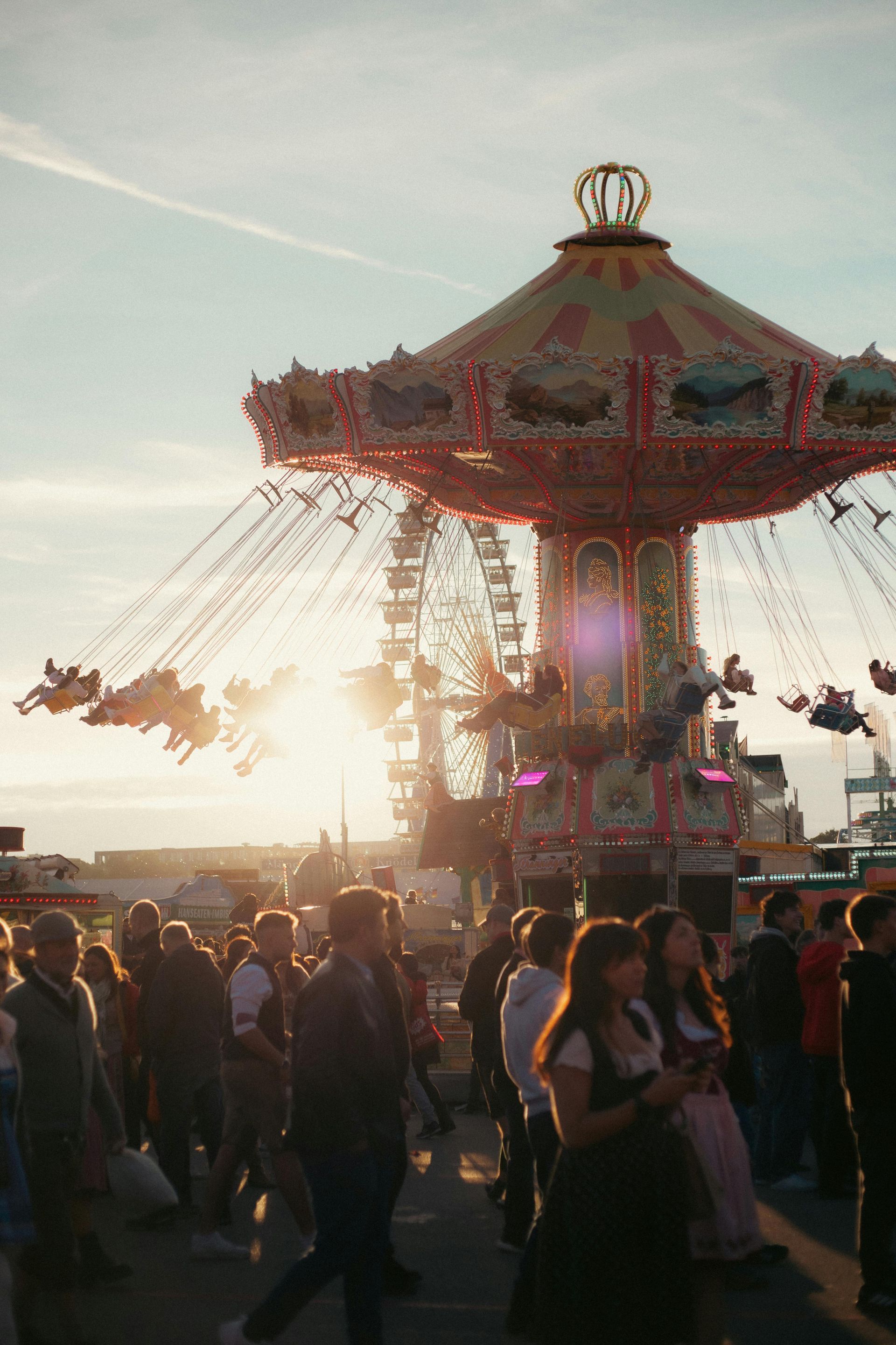 Fairground scene with a carousel and Ferris wheel at sunset. Crowd of people.