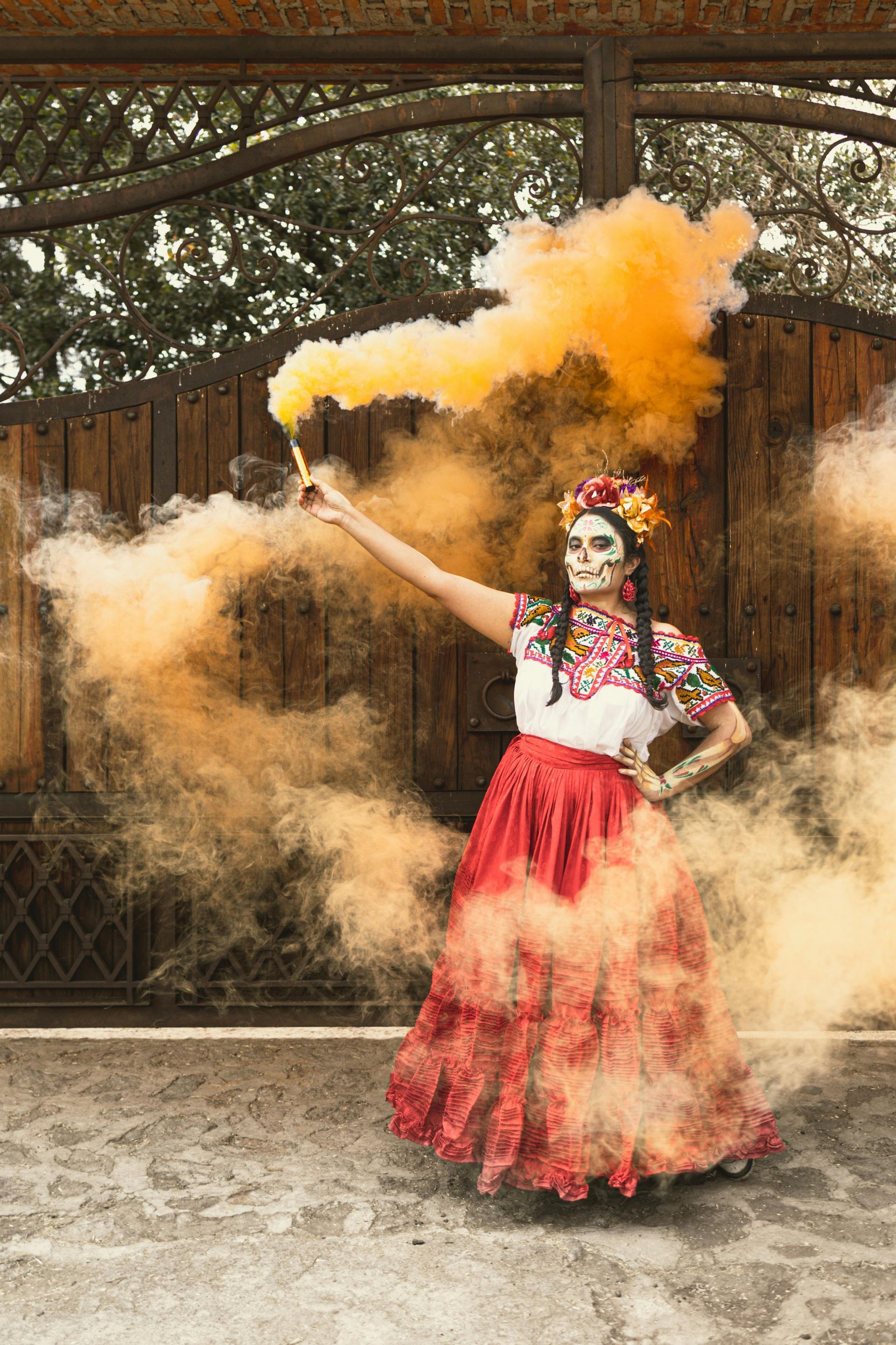 Woman in Day of the Dead attire holds orange smoke flare, standing by a wooden gate, outdoor setting.