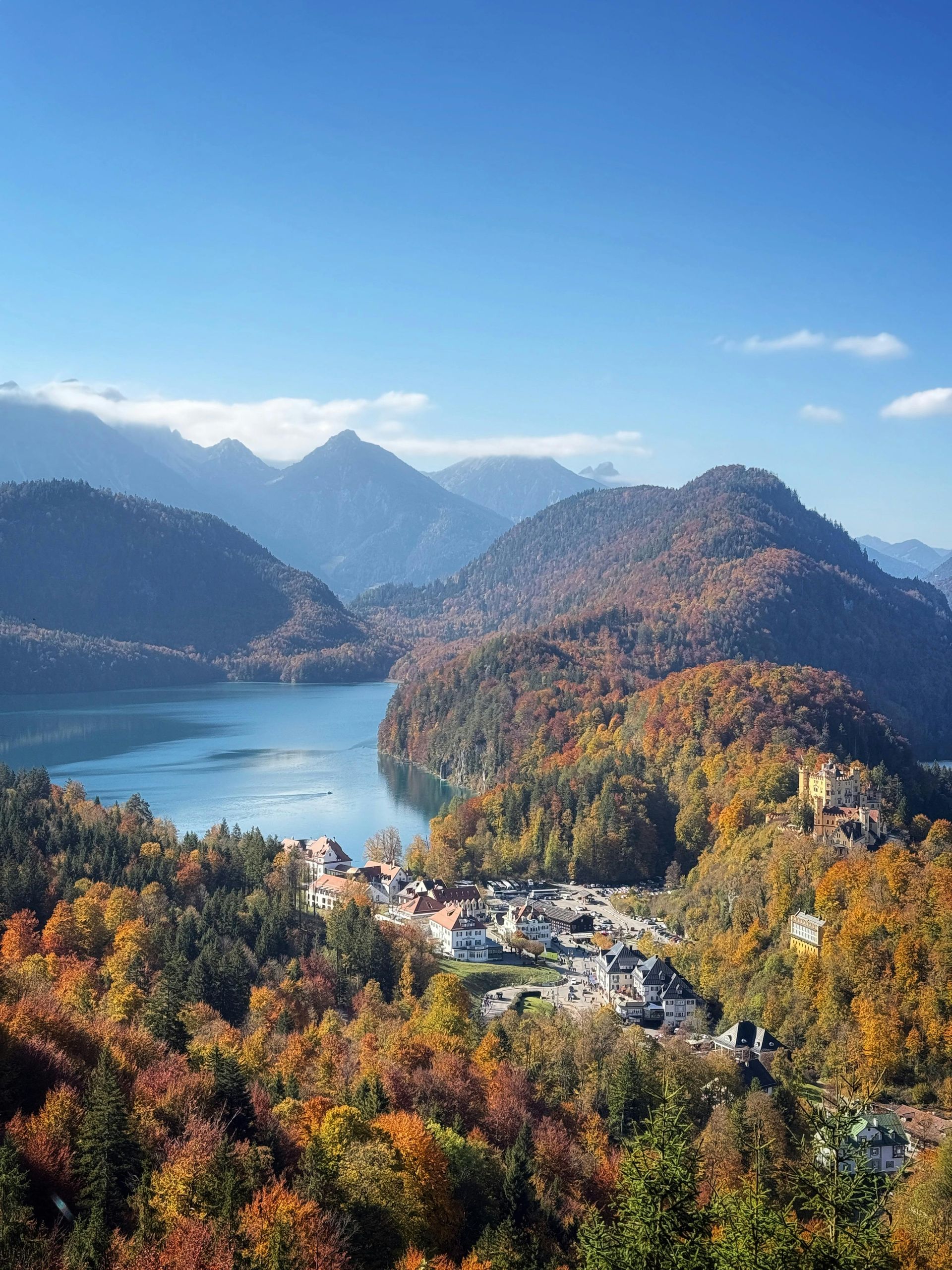 View of a lake and town nestled between autumn-colored hills and mountains under a blue sky.