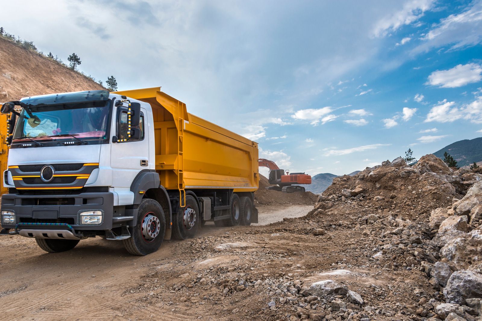 A dump truck is driving down a dirt road next to a pile of rocks.
