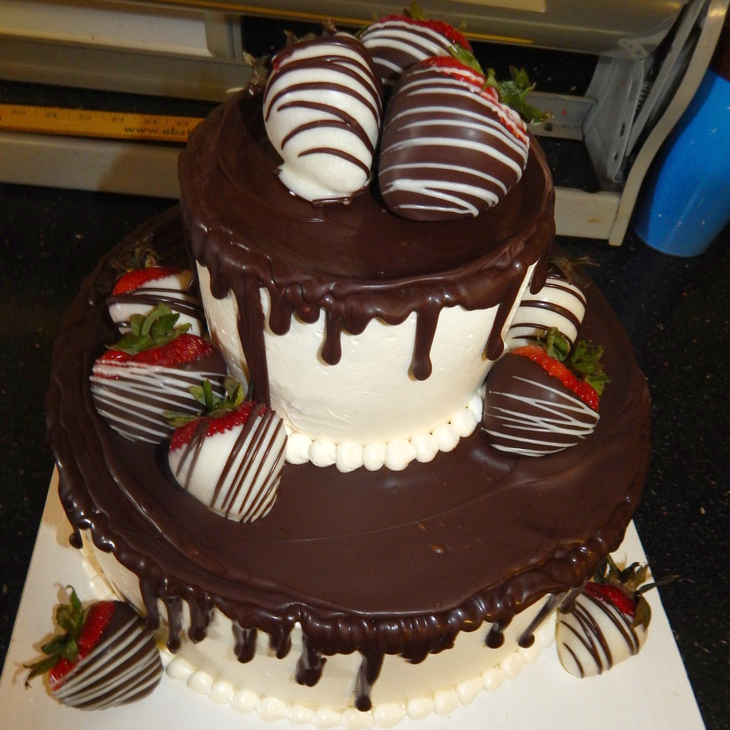White tiered wedding cake with dark chocolate dripped over the top, and decorated with chocolate dipped strawberries