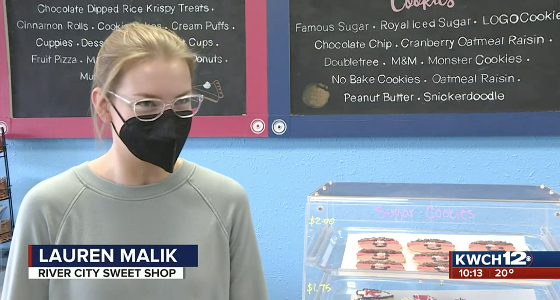Blond woman in a mask standing in front of a glass case full of cookies.