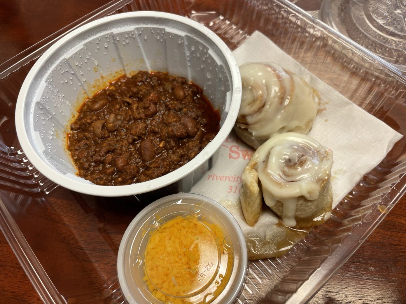 A cup of chili, two cinnamon rolls, and a cup of cheese in a to-go box