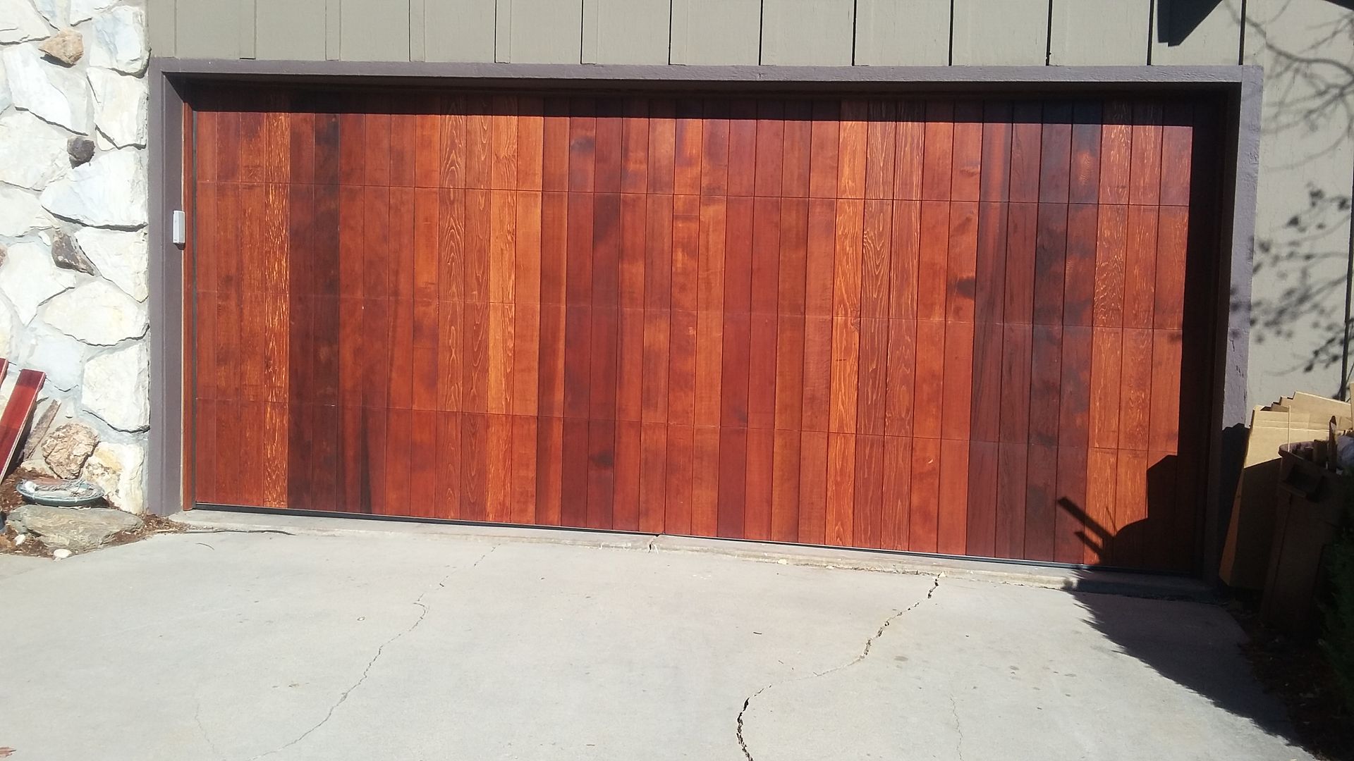 Wooden garage door with vertical planks, brown hues, set in concrete, next to a stone wall.