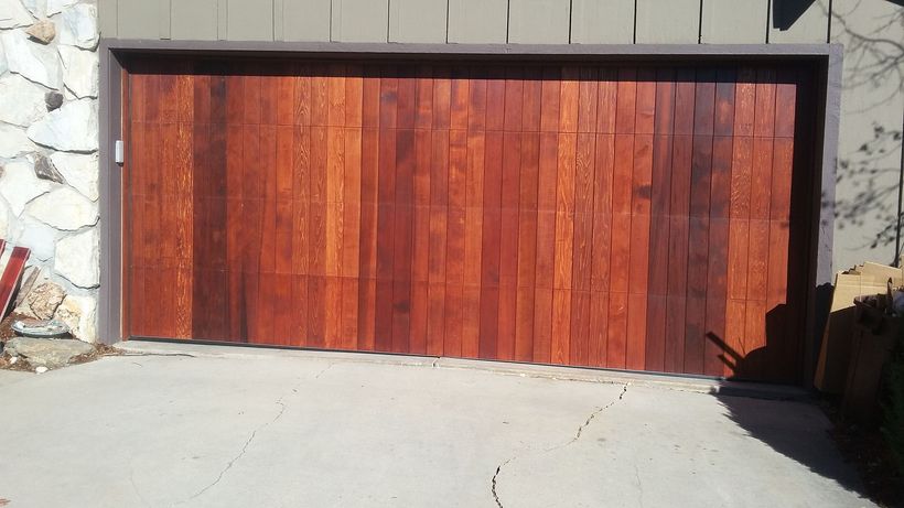 Wooden garage door with vertical planks, brown hues, set in concrete, next to a stone wall.