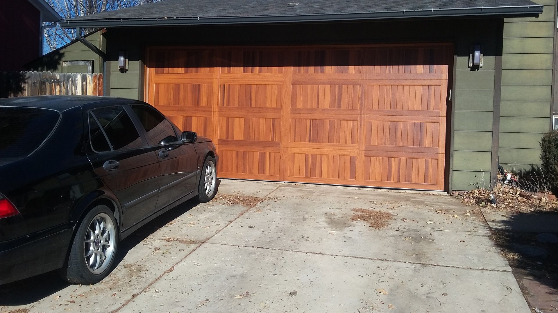 Black car parked in front of a closed, brown wooden garage door in a driveway. Sunny day.