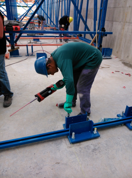 Obrero de la construcción aplicando sellador a una estructura metálica azul sobre un suelo de hormigón.