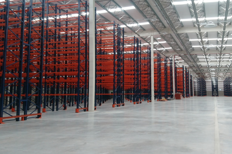 Almacén vacío con altas estanterías industriales naranjas y azules. Suelo de hormigón y techo metálico.
