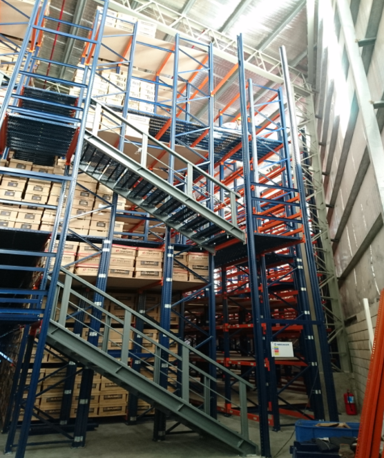 Estanterías de almacén multinivel con escaleras y cajas, vigas azules y naranjas.