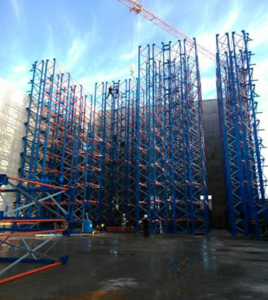 Estructura de estanterías industriales azules y naranjas en construcción; trabajadores presentes.