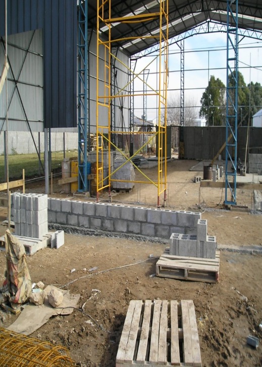 Obra en construcción con muro de bloques de hormigón en construcción, andamios y almacén al fondo.