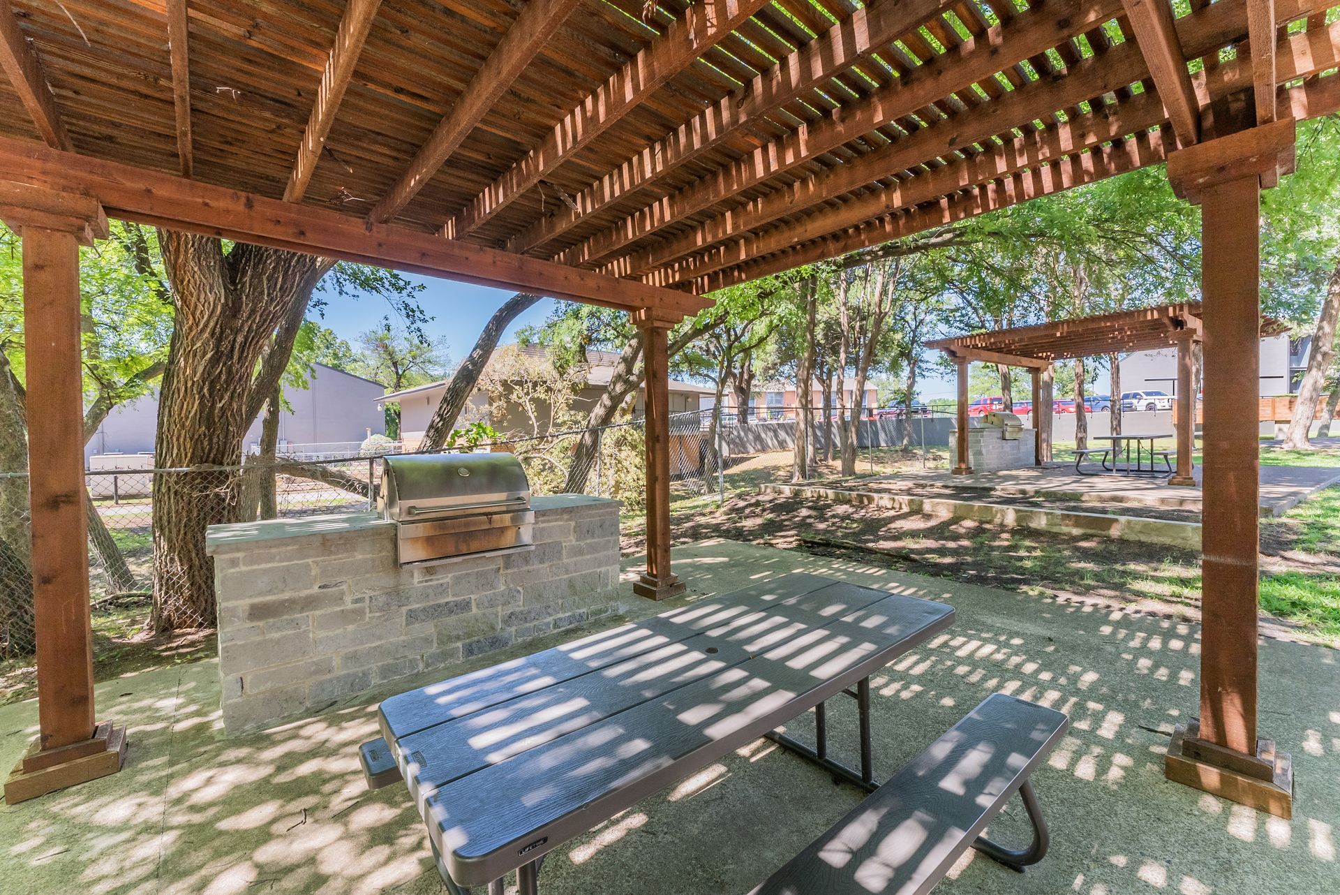 A picnic table under a wooden pergola with a grill.