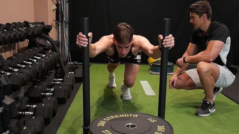 Man pushing weighted sled; trainer watches in gym.