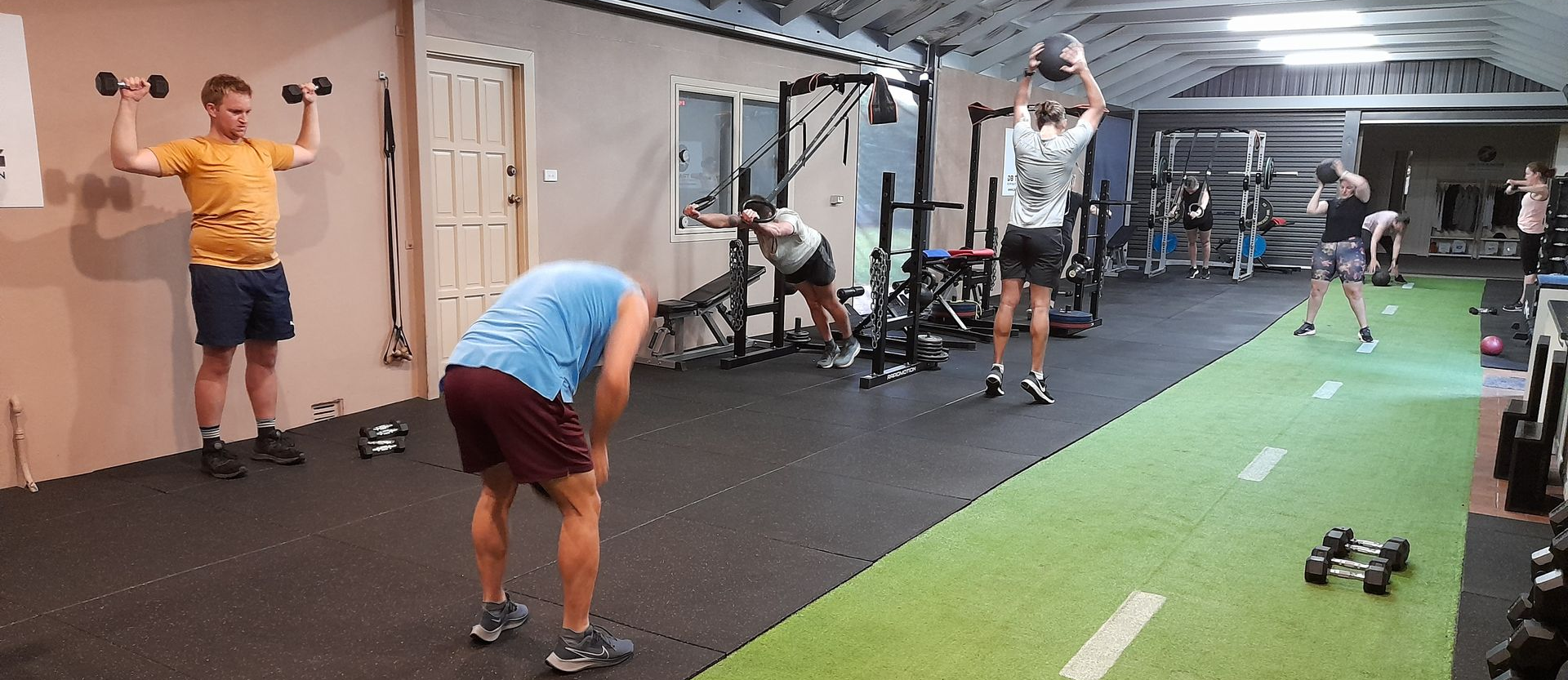 People exercise in a gym. One man lifts dumbbells. Others use a medicine ball and work out.