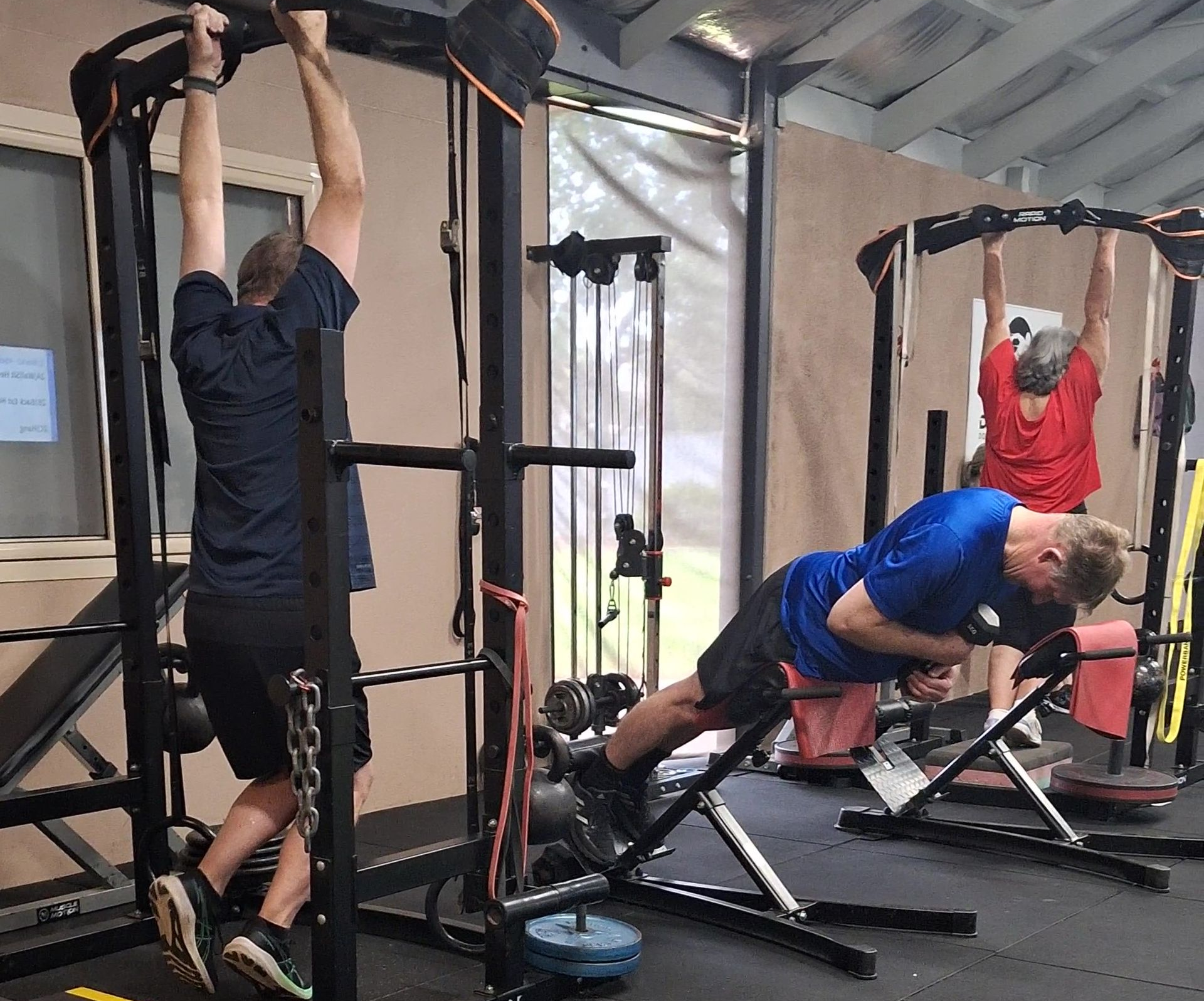 People exercising in a gym; one doing pull-ups, another a back extension, and a third hanging from a bar.