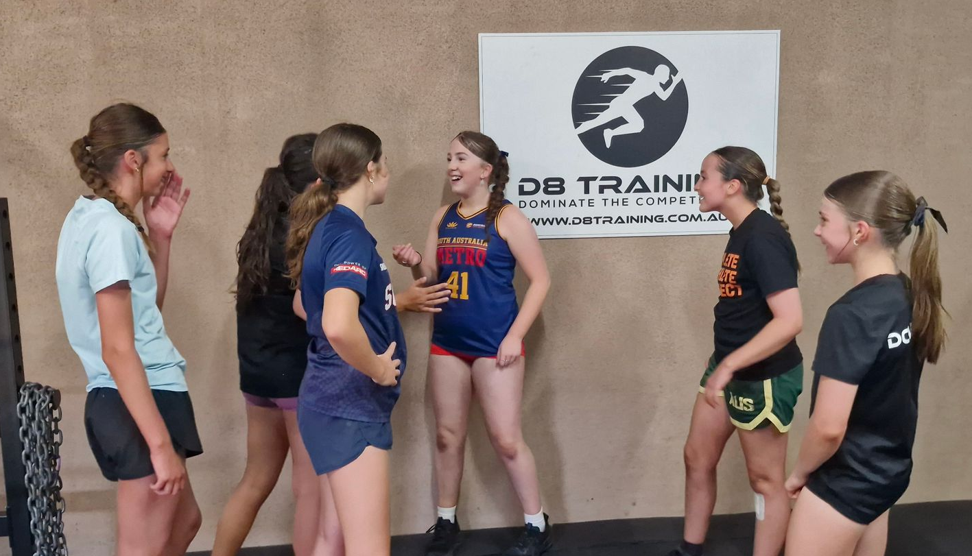 A group of girls in athletic wear laughing in a gym with a training logo on the wall.
