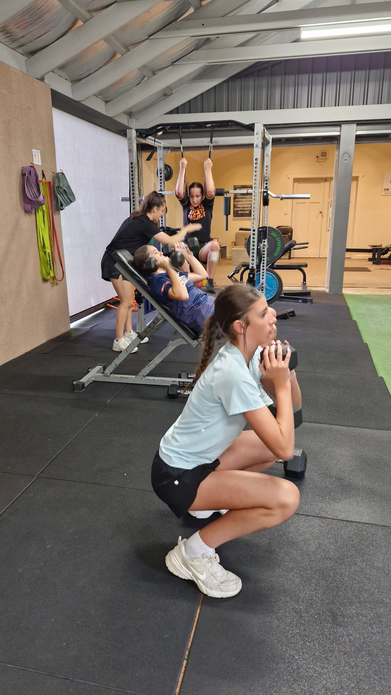 Woman squats with dumbbell in gym; others do exercises in background.