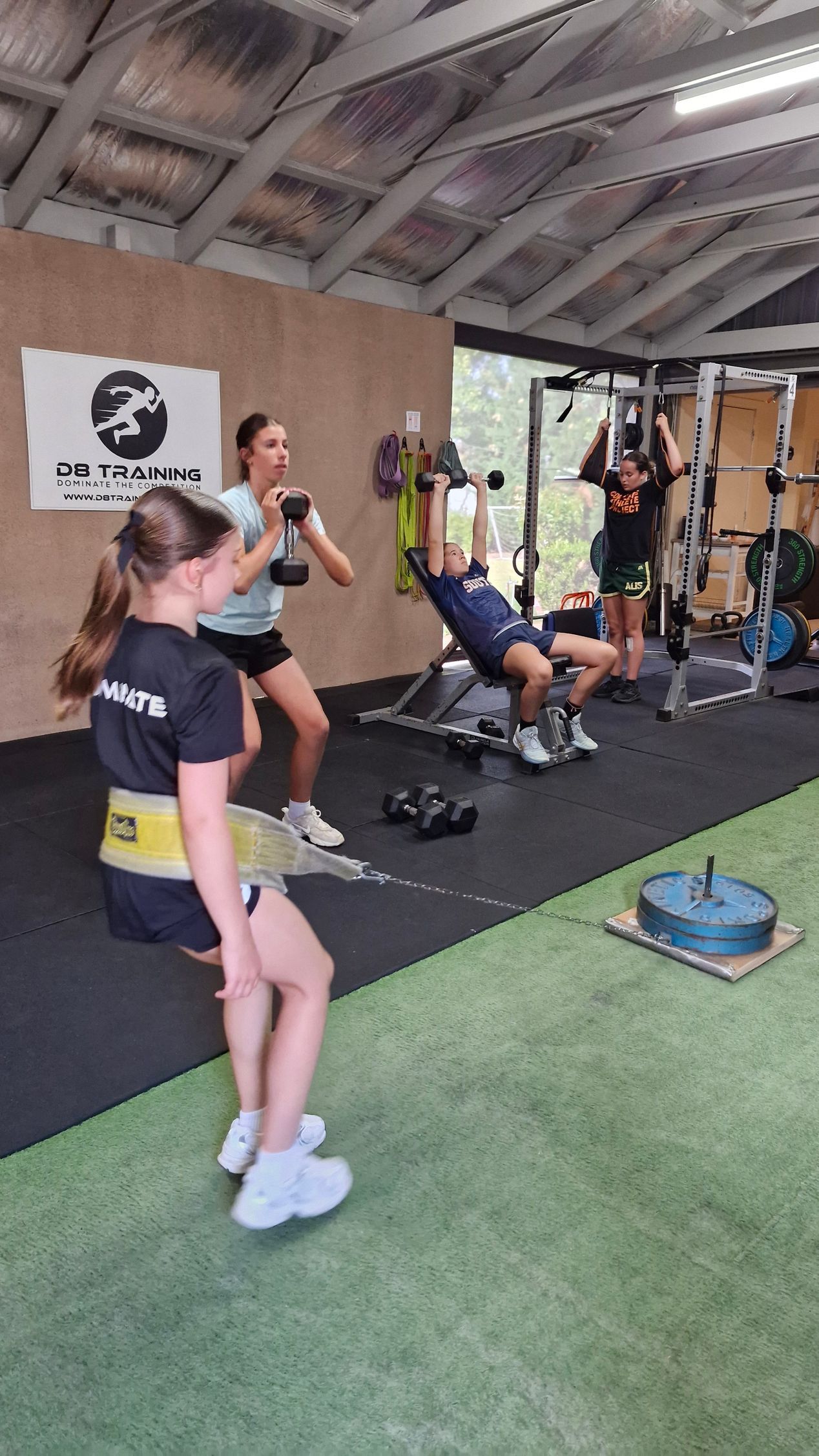 People exercising in a gym. One is doing a chest press, others are using dumbbells. A logo is visible on the wall.