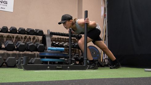 A person pushes a weighted sled across a gym floor, arms extended, head down.