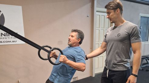Man performing ring rows with trainer's assistance in gym.