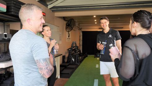 Four people in gym, chatting and holding water bottles. One person wears a black polo, the others are in activewear.