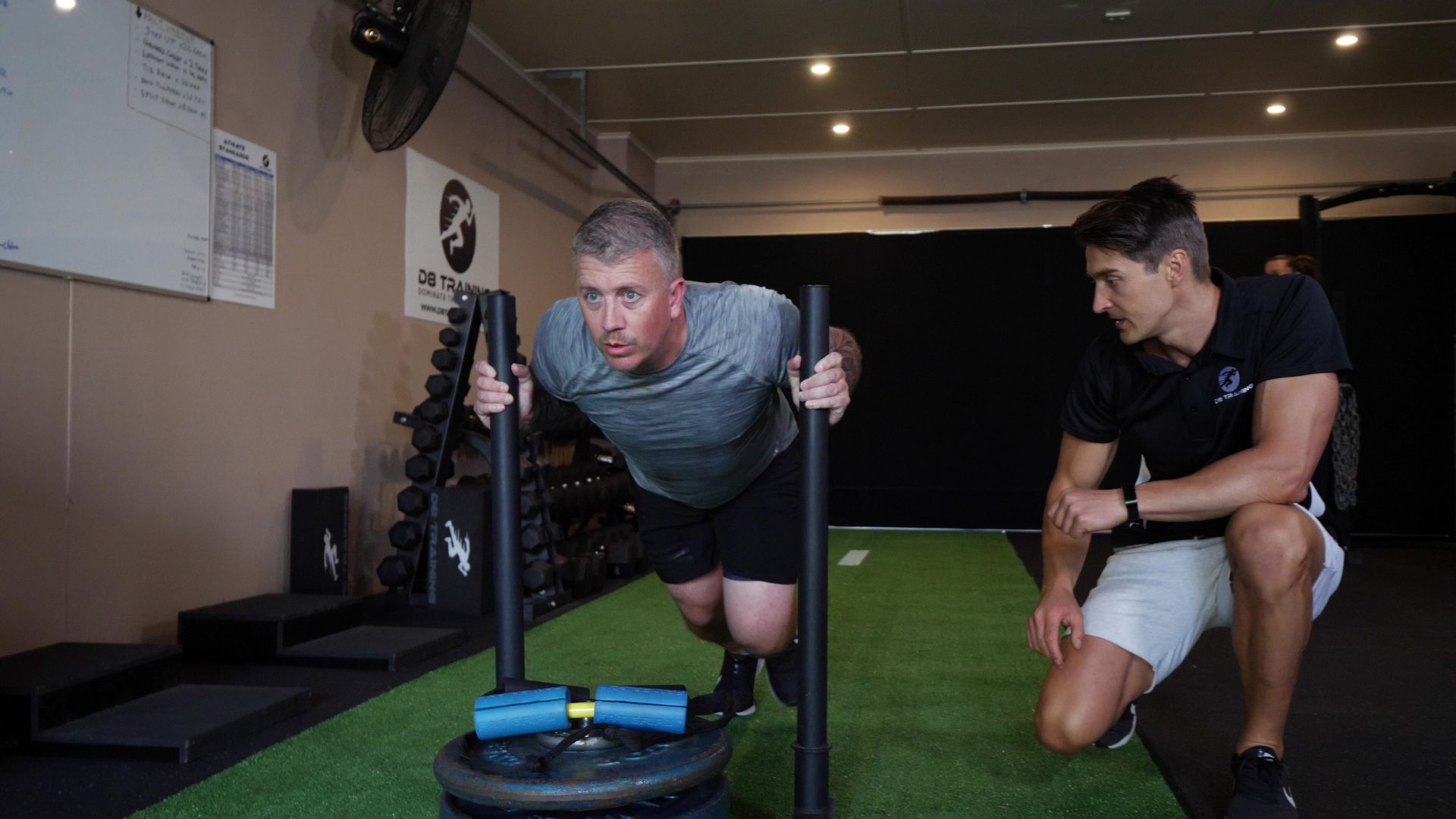Man pushing a weighted sled, supervised by a trainer, in a gym.