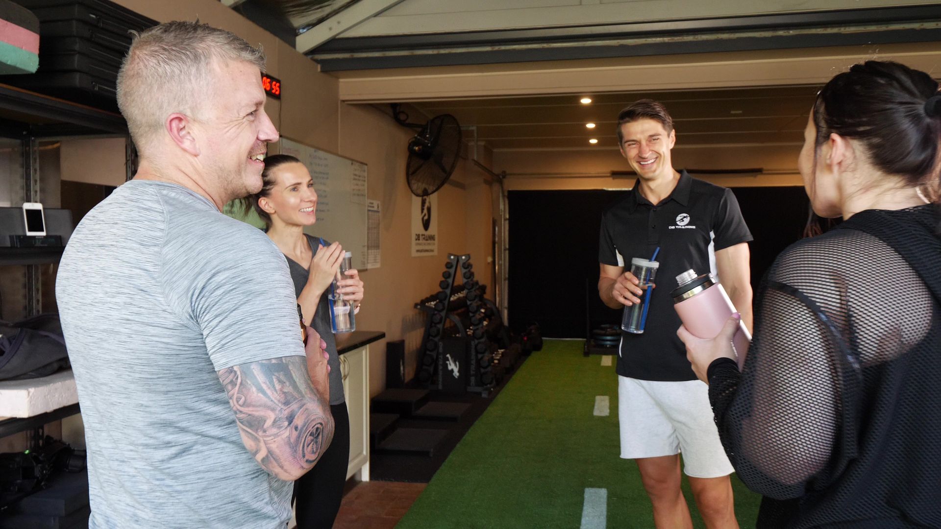 People in a gym chat, holding water bottles. A trainer smiles, and there is gym equipment in the background.