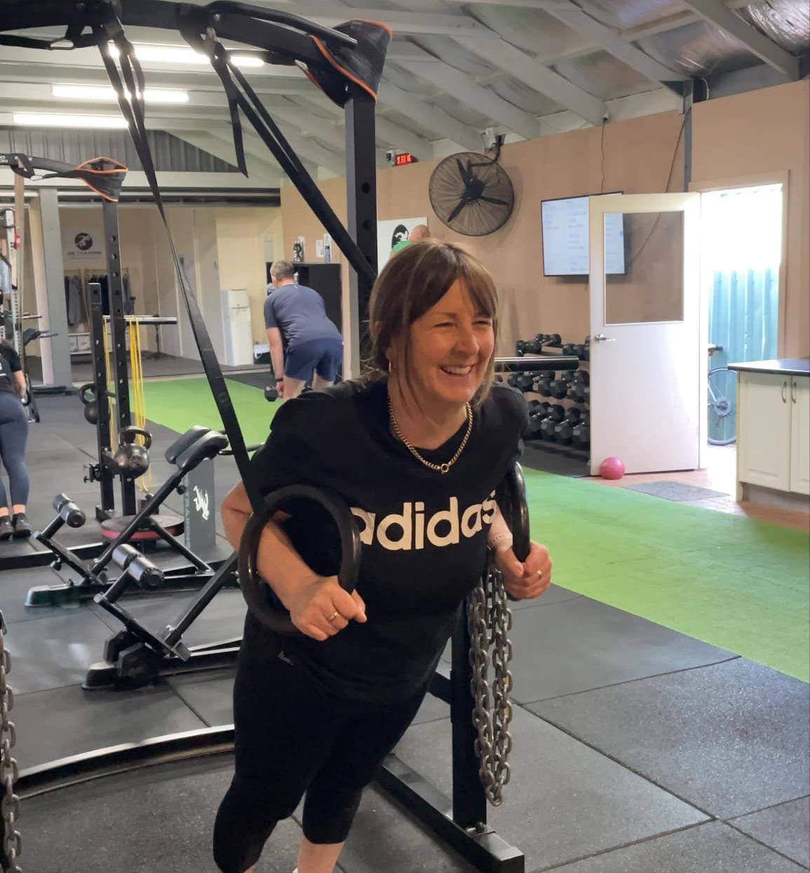 Woman using gymnastic rings in a gym, smiling. Black Adidas shirt, green floor, others exercising in background.