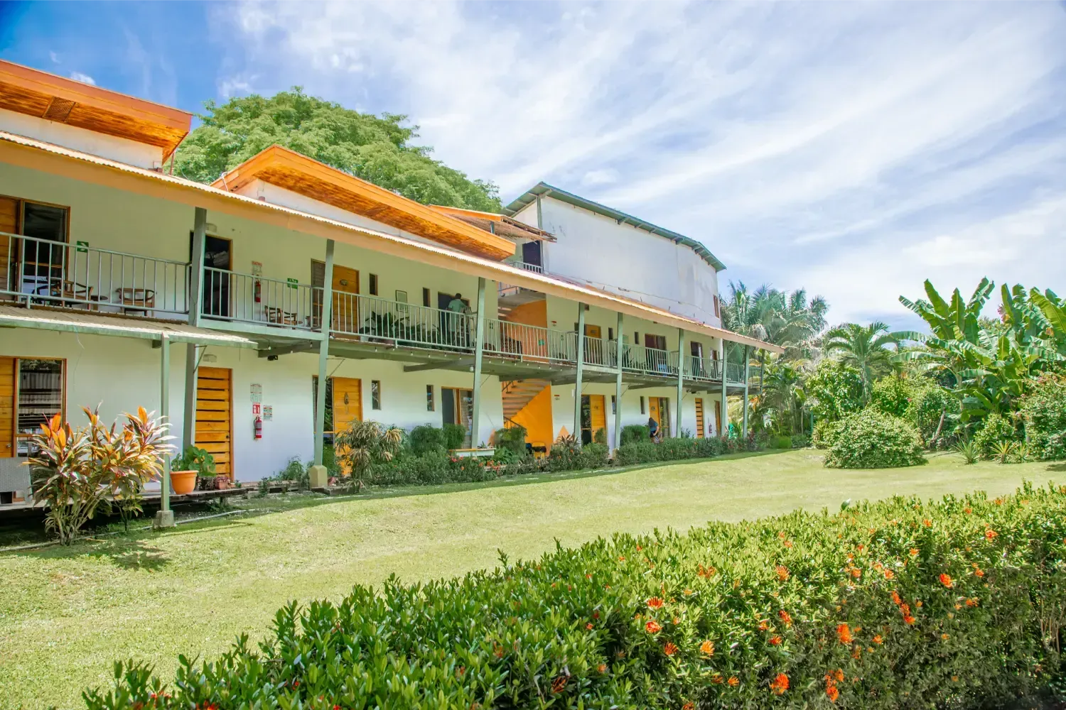 Two-story white building with balconies, yellow doors, and green lawn, with tropical plants under a blue sky.