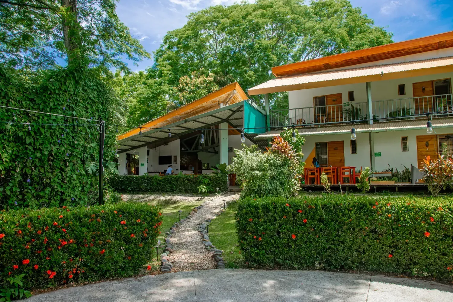 White building with orange roof in a lush, green tropical setting, featuring a stone walkway and manicured hedges.